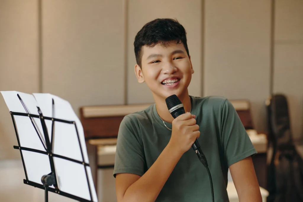 Smiling teenage boy holding a microphone beside a music stand during a singing lesson.
