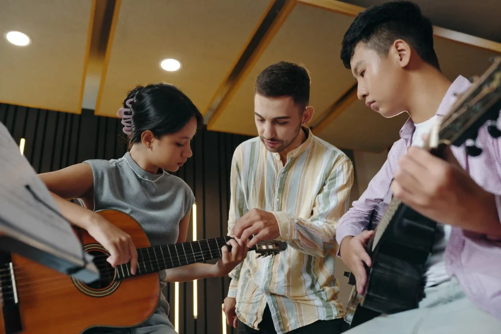 Instructor guiding a woman’s finger placement on an acoustic guitar while another student practises during group guitar lessons.
